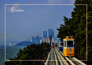 Die erhöhte Bahnstrecke führt durch viel Grün und zieht den Blick auf die Küste und die entfernte Skyline von Busan, während die gelbe Bahn als auffälliger Farbakzent durch die Landschaft fährt.