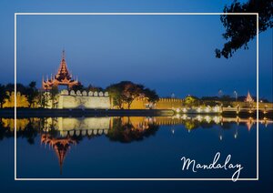 Entlang des Wassers leuchten die Mauern der Tempelanlage in Mandalay im warmen Licht, während sich die Gebäude klar im ruhigen See spiegeln. Der Himmel darüber wirkt tiefblau und bildet einen starken Kontrast zu den goldenen Details und den beleuchteten Uferlinien.