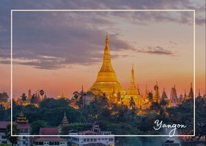Der Shwedagon-Pagoda-Tempel erhebt sich golden über die umliegende Stadt und leuchtet im warmen Licht des Sonnenuntergangs. Rundherum verdichtet sich das Stadtbild mit vielen kleineren Gebäuden und einer ruhigen Abendstimmung im Himmel. Er sieht von weitem fast wie der Pariser Eifeturm aus, mit seiner hell scheinenden Beleuchtung und der spitzen Form.
