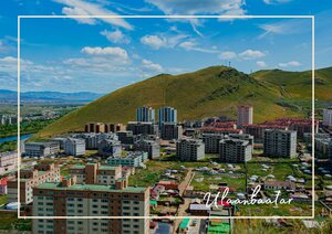 Die Hauptstadt der Mongolei, Ulaanbaatar, liegt mit ihren dicht stehenden Gebäuden vor einem grünen Hügel. Zwischen den modernen Häusern und den offenen Hängen entsteht ein deutlicher Kontrast zwischen Stadt und Landschaft. Über der Stadt öffnet sich ein weiter Himmel.