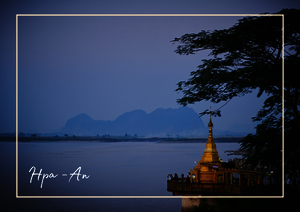 Der Hpa-An-Ort liegt ruhig am Wasser, während ein goldener Tempelstupa auf einer kleinen Plattform am Ufer sichtbar ist. Im Hintergrund zeichnen sich sanfte Bergsilhouetten ab, und die spiegelnde Wasserfläche betont die offene Landschaft.