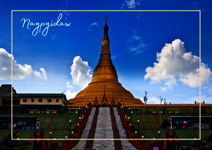 Vor einem klaren Himmel führt ein gerader Weg direkt auf die goldene Shwedagon-Pagode zu. Die Tempelanlage wird von niedrigen Gebäuden und offenen Flächen eingerahmt, wodurch der hohe Stupa besonders deutlich hervorsticht.