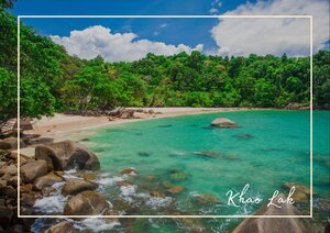 In Khao Lak schimmert das Meer in klarem Türkis und legt sich sanft an den hellen Sandstrand, während glatte Felsen den Uferbogen rahmen. Das dichte Grün der Hügel wächst bis dicht ans Wasser heran und gibt der Bucht etwas Frisches und Tropisches. Über allem spannt sich ein weiter blauer Himmel mit weißen Wolken, der die Szene licht, ruhig und einladend wirken lässt.