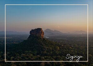 Der Sigiriya Rock hebt sich dunkel und massiv aus einer weitläufigen, grünen Landschaft hervor, die im sanften Licht in verschiedenen Schattierungen von Grün bis Goldbraun leuchtet. Über dem Felsen liegt ein heller Himmel mit zarten Orange- und Blautönen, während die Umgebung still und weit wirkt und dem Ort eine besondere Größe verleiht.
